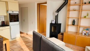 A compact living space with a kitchenette, a sofa, a wooden cabinet with shelves, and a black wood-burning stove. - Ai Ronchi by Interhome (Gordola)