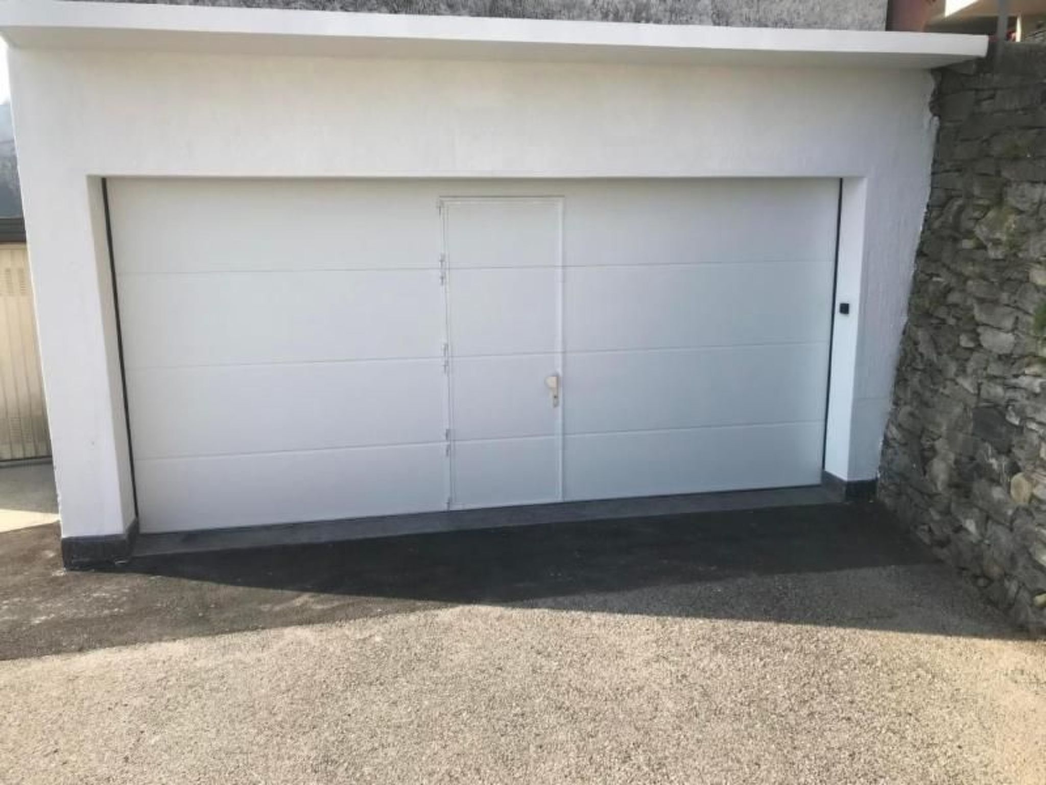 Un garage moderne, à l’extérieur épuré et blanc, avec un mur en pierre sur le côté droit.