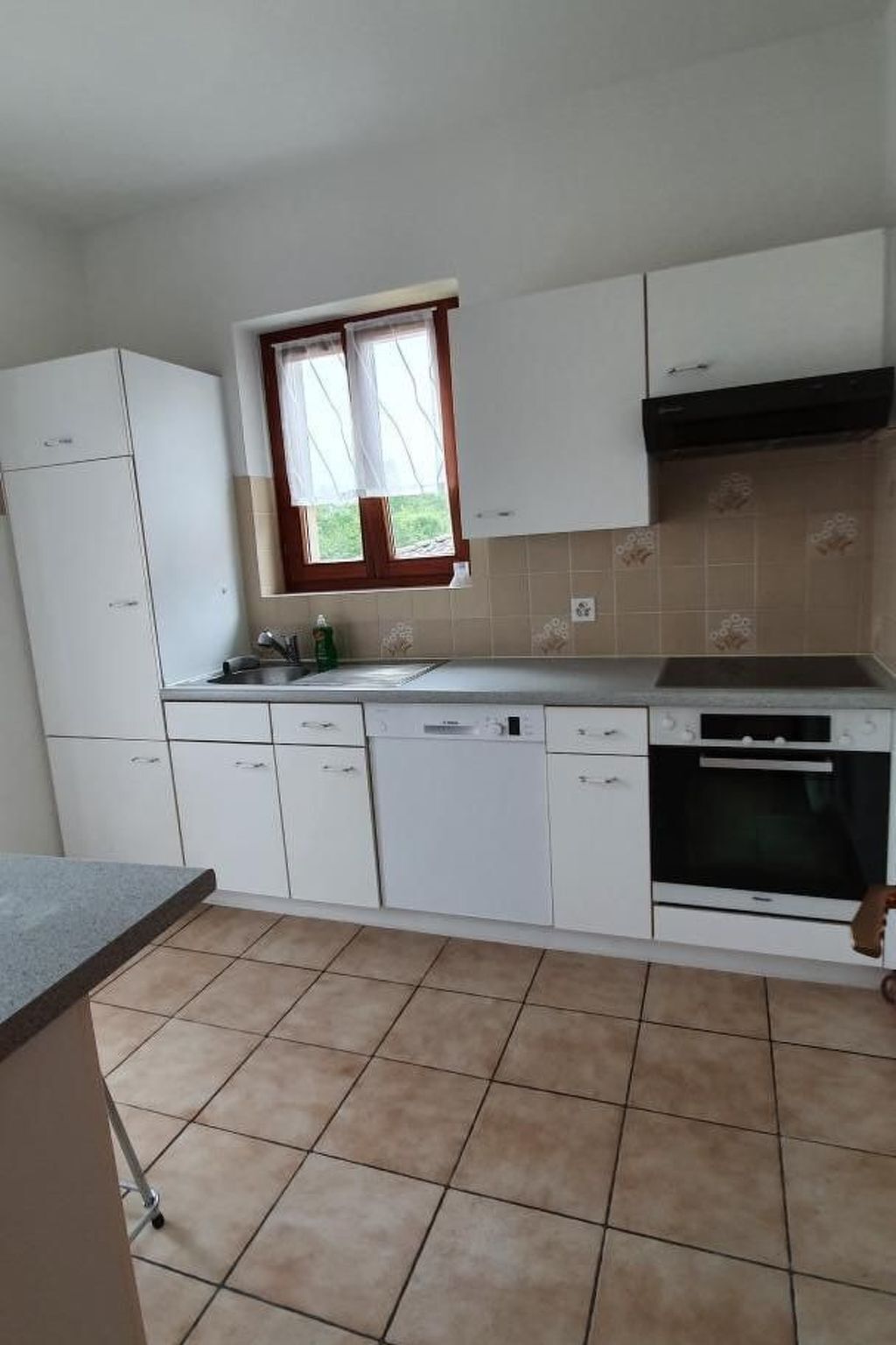 Eine Küche mit weißen Schränken, einem Geschirrspüler, einem Backofen und einem Fenster mit Vorhang.
