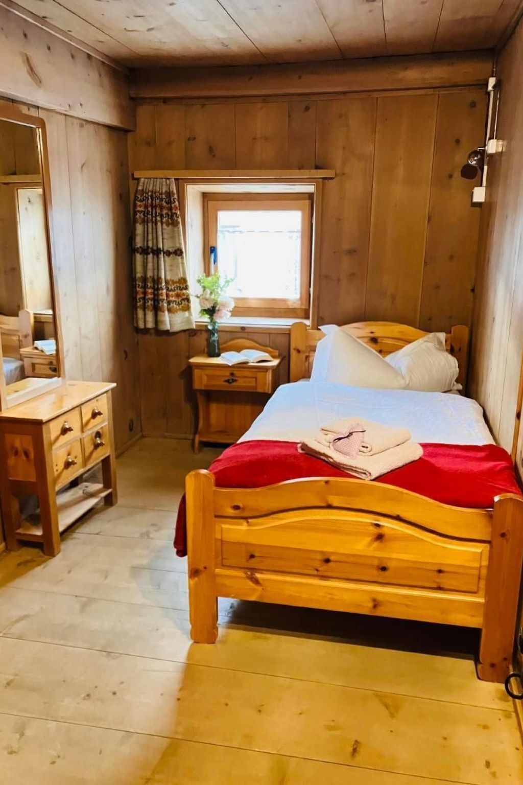 Une chambre dans une cabane en bois, avec un lit simple, une table de chevet, une commode et une fenêtre avec des rideaux.