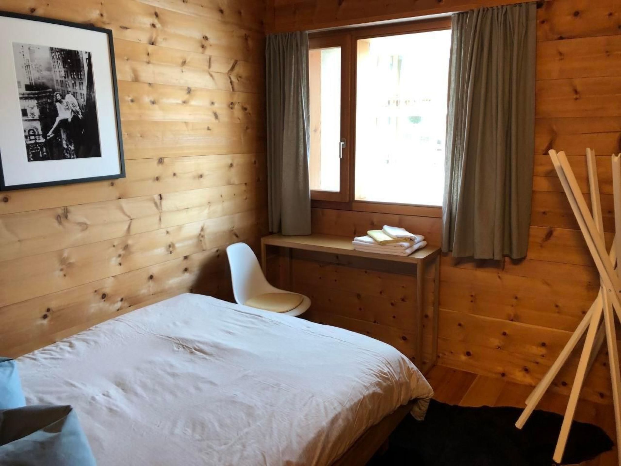 A wooden cabin room with a bed, a desk, and a chair. There is a window with curtains, a framed picture on the wall, and a black rug on the floor.
