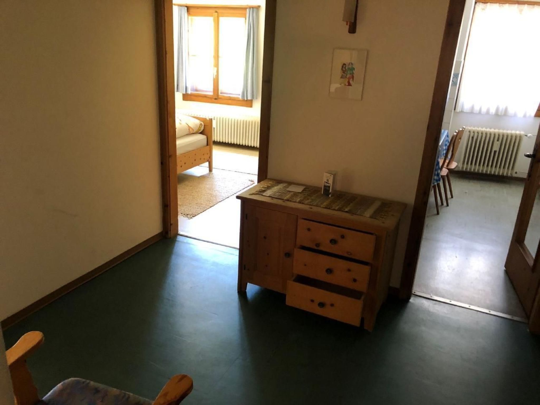 A small room with a wooden cabinet, a chair, and a view of a bed and radiator through an open door.