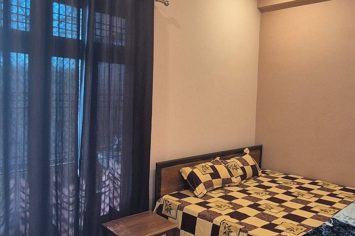 Ein Schlafzimmer mit einem Bett, Deckenventilator, einem Fenster mit Vorhängen, einem kleinen Tisch und einem Fliesenboden.