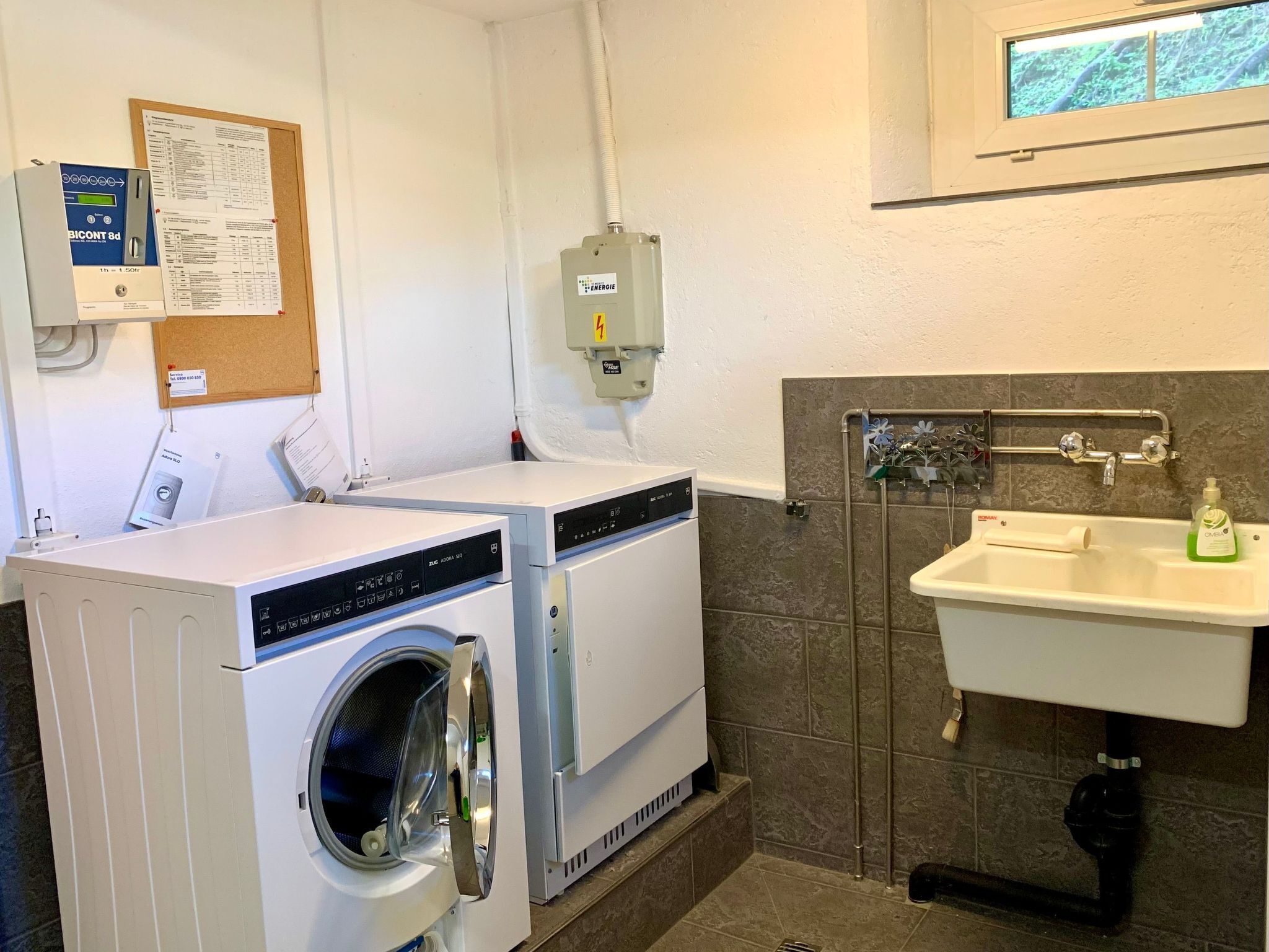 A laundry room with two front-loading washing machines, a utility room with a water heater, and a sink with a hand soap dispenser.