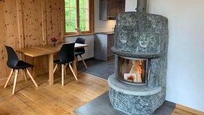 Ein gemütliches Zimmer mit Kamin aus Stein, Holzwänden, einem Esstisch mit Stühlen und einem Fenster mit Blick auf Grünflächen.