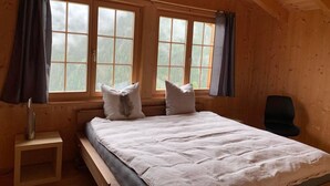 Ein Schlafzimmer in einer Holzhütte mit einem Bett, zwei Fenstern, einem Sessel und einem kleinen Tisch.