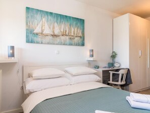 Een slaapkamer met een bed, een bureau, een stoel, een kledingkast en een schilderij van zeilboten aan de muur.