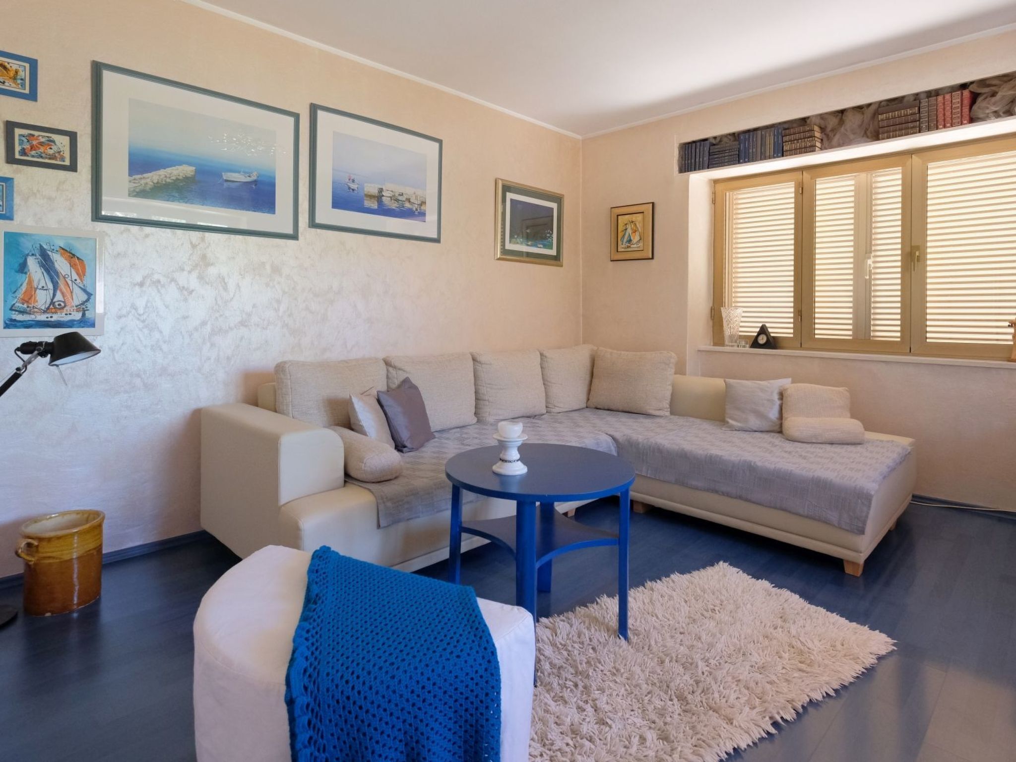 A living room with a beige sofa, a blue table, and a white rug. There are framed pictures on the wall and a window with blinds.