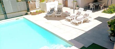 Ein Pool mit Liegestühlen und einem Tisch, umgeben von einer Terrasse mit Pflanzen und einem kleinen Gebäude.