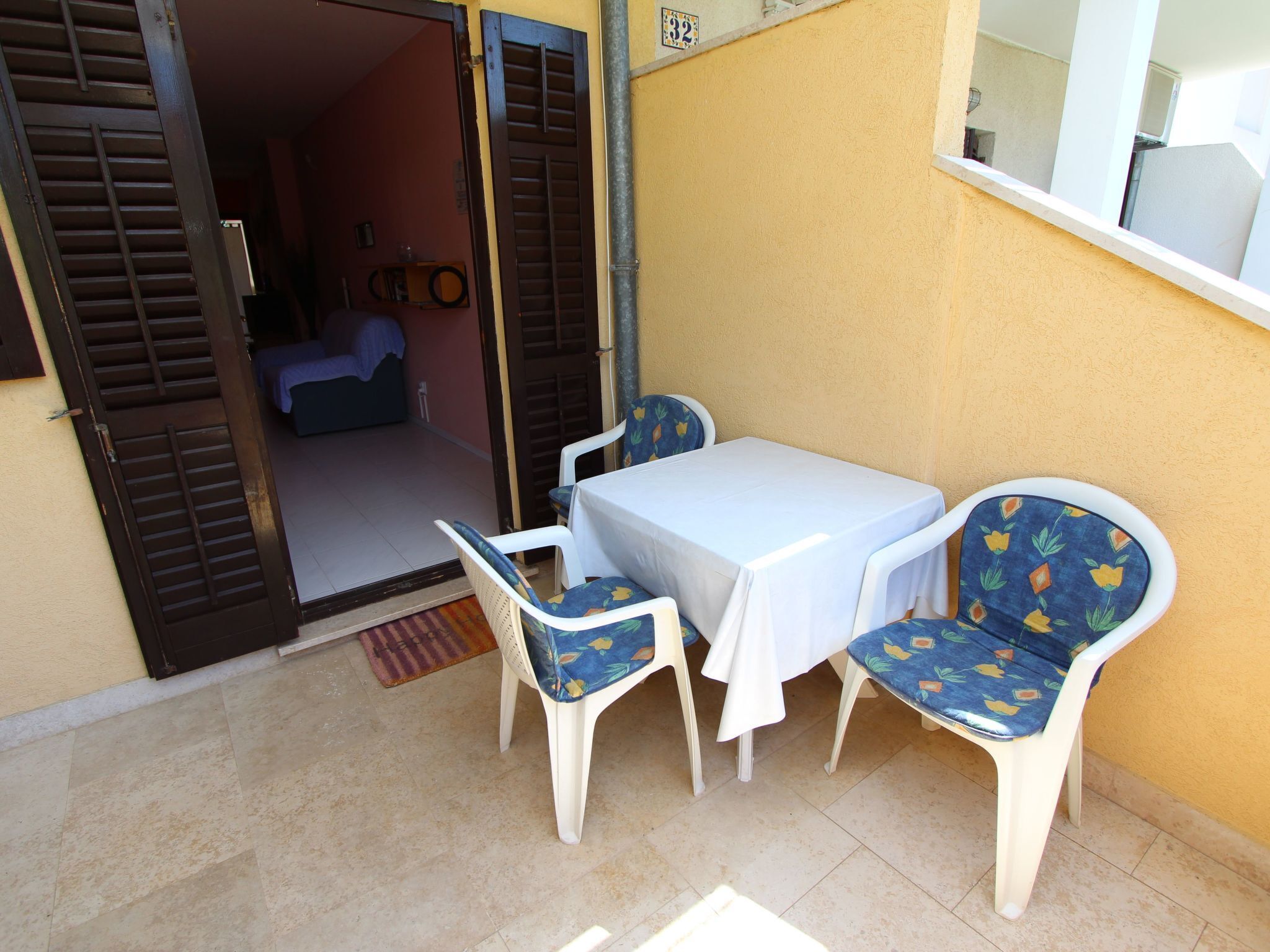 A small outdoor seating area with a table and two chairs, a door leading to a room, and a window with a view.