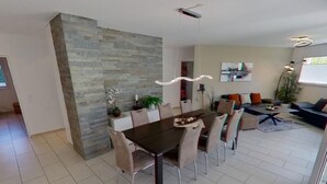 Ein moderner Essbereich mit einem dunklen Holztisch und beigen Stühlen, einer Wand mit Steinakzenten und Blick auf einen Wohnbereich mit einer Couch und einer Pflanze.
