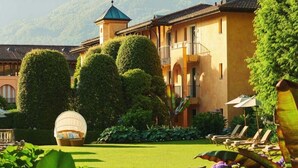 A hotel with a well-manicured garden, a pool area with lounge chairs, and a traditional building with a tower. - Maisonettewohnung in Residenz des Giardino Ascona by Interhome (Ascona)