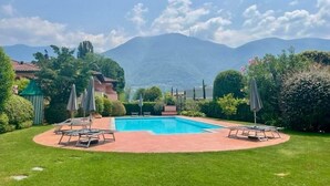 A pool with lounge chairs and umbrellas, surrounded by well-maintained grass and hedges, with mountains in the background. - Maisonettewohnung in Residenz des Giardino Ascona by Interhome (Ascona)