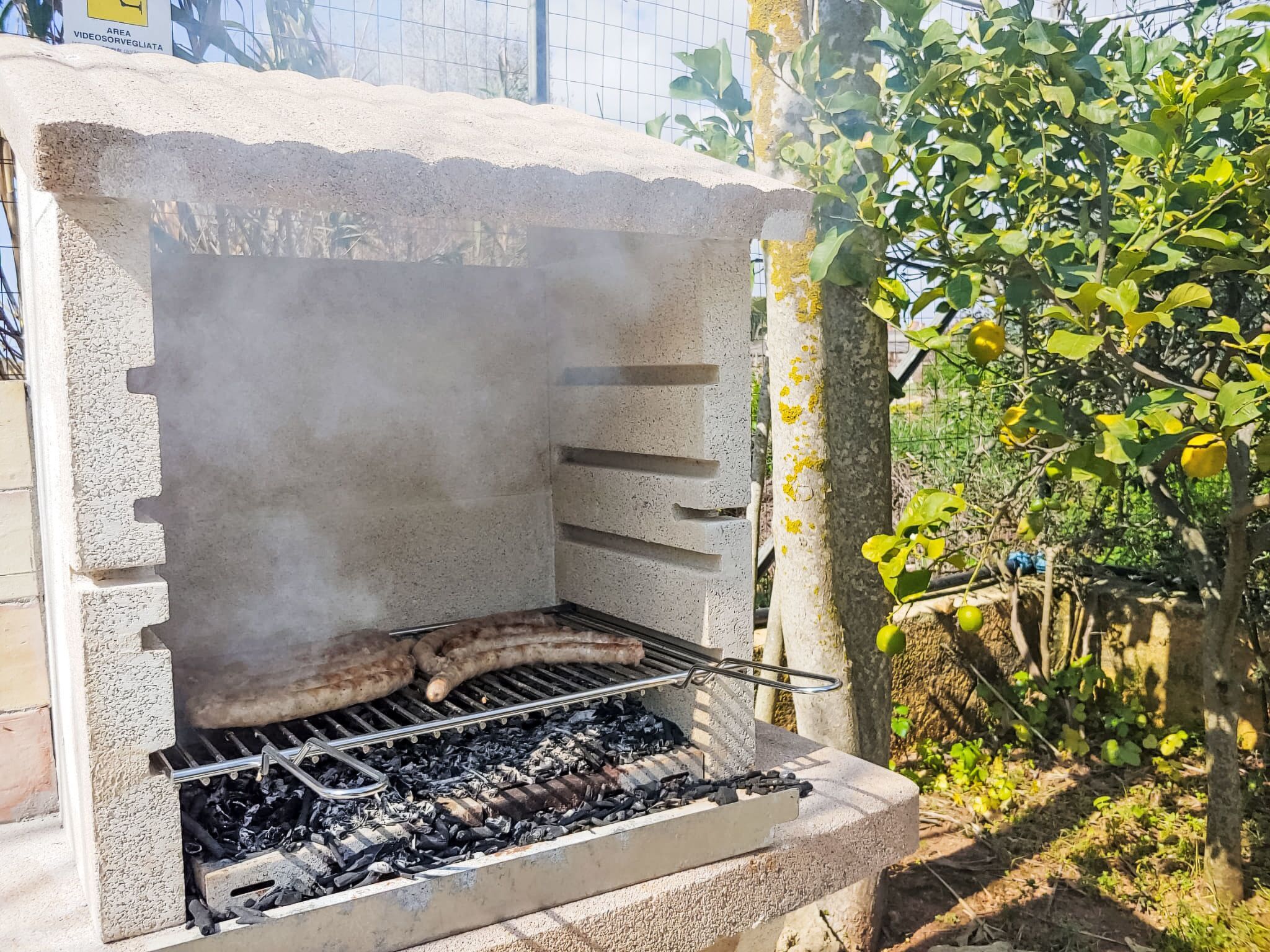 Una griglia per barbecue in cemento con una griglia metallica, immersa nel verde e delimitata da una recinzione.