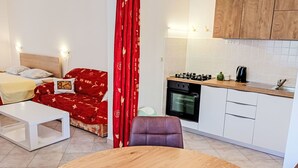Ett kompakt bostadsutrymme med en säng, en röd, mönstrad fåtölj, ett matbord, ett köksområde med spis och ugn samt vita skåp.