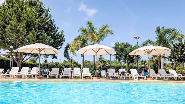 Una piscina con sillas de descanso y sombrillas, rodeada de palmeras y otra vegetación.