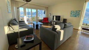 Una sala de estar moderna con dos sofás grises, un sillón rojo, un televisor y un ventanal con vistas a la montaña.