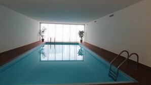 Una piscina interior con agua azul cristalina, suelo de baldosas y vistas al exterior a través de amplias ventanas.