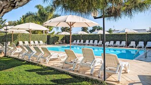 Ein Poolbereich mit Liegestühlen und Sonnenschirmen, ein klares, blaues Schwimmbecken und ein Heckenstreifen im Hintergrund.