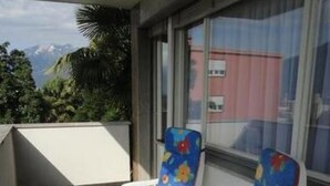 Un balcone con due sedie bianche rivestite con motivi floreali colorati, un pavimento a righe e vista su vegetazione e un edificio sullo sfondo.