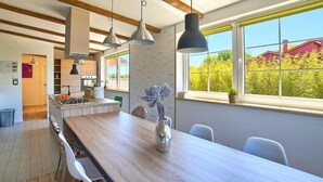 Een moderne keuken met houten balken, hanglampen, een eethoek met een lange tafel en uitzicht op groen.