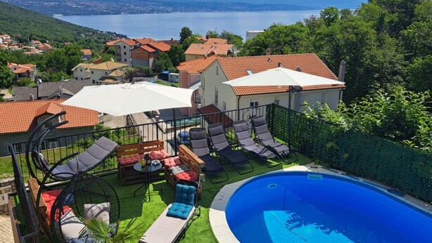 Area piscina con lettini e vasca idromassaggio, circondata da recinzione e vegetazione, con vista su un lago e case sullo sfondo.