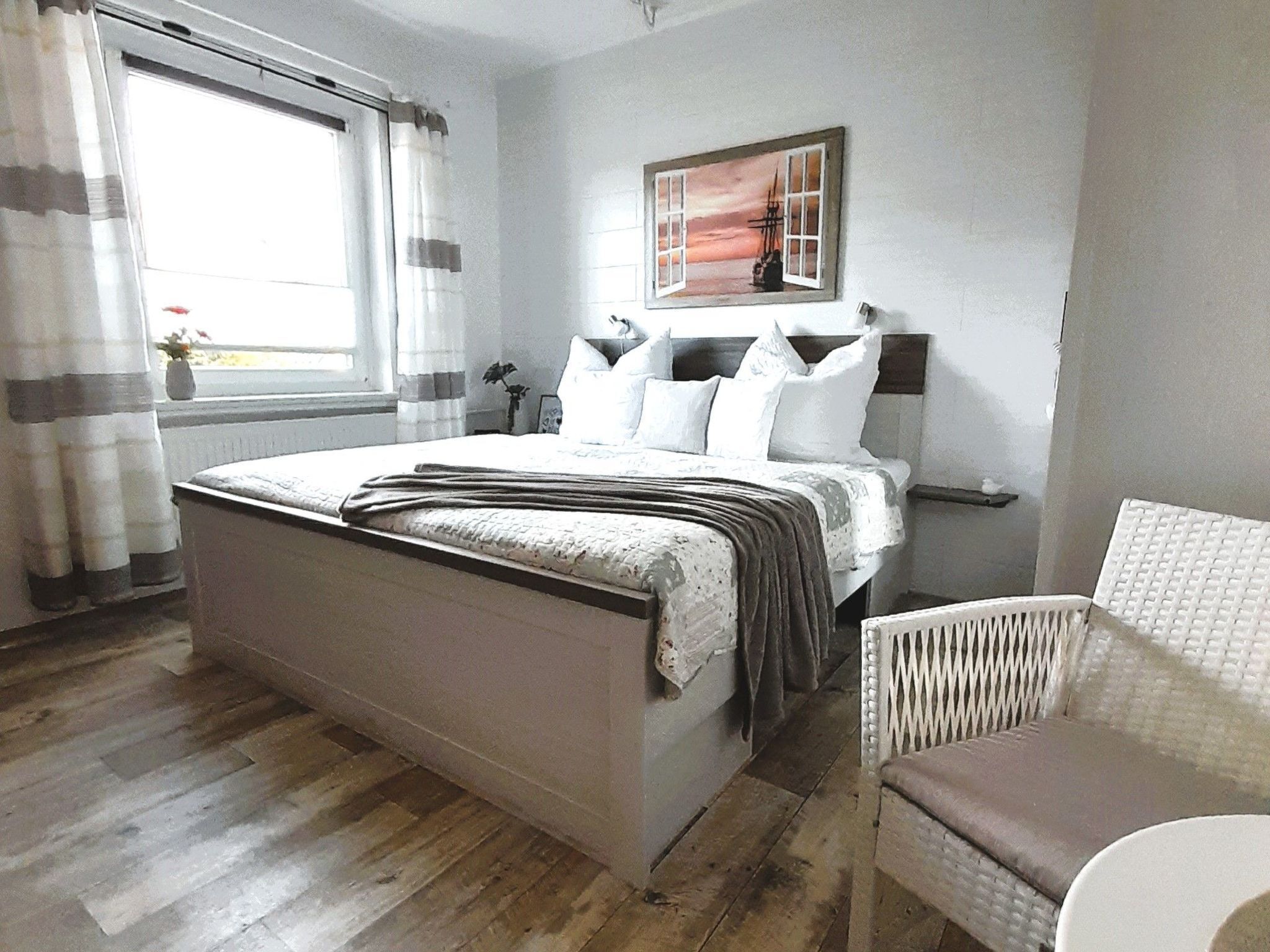 Une chambre avec un grand lit, une chaise, un tableau au mur et une fenêtre avec des rideaux.
