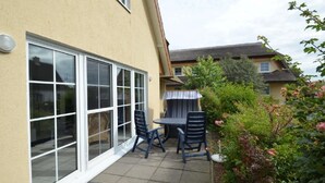 Un patio con mesa y sillas, una puerta corrediza de vidrio y un jardín con arbustos y flores.