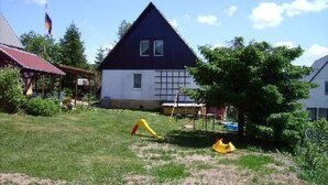 Ein Haus mit dunkelblauem Dach und weißen Wänden, ein kleiner Garten mit einer gelben Rutsche und eine Flagge an einem Mast.