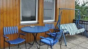 Eine überdachte Terrasse mit einem runden Tisch und blauen Stühlen, einem Schaukelstuhl und zwei Fenstern mit weißen Rahmen.