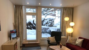 Un salon chaleureux avec un canapé rouge, une table basse en bois, un fauteuil noir et un téléviseur posé sur un meuble. De grandes fenêtres offrent une vue sur un paysage enneigé.