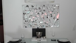 Un espace de salle à manger moderne avec une table en verre, des chaises blanches, une grande œuvre d’art murale en métal et un luminaire suspendu.