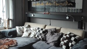 Une chambre moderne avec un canapé gris, des coussins à carreaux et une étagère murale décorée d’œuvres d’art.