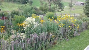 Un paysage rural avec une clôture en bois, différentes plantes en fleurs et une grange visible au loin.