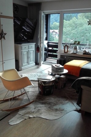 Un salon moderne avec un piano, un canapé, une table basse et une vue sur un balcon doté d’une balustrade et de plantes vertes.