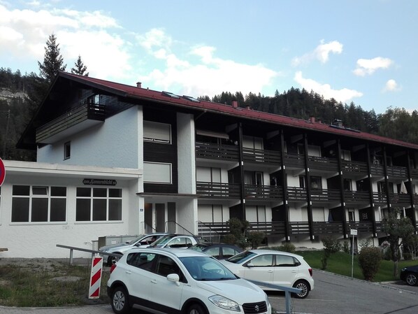 Un immeuble à plusieurs étages doté de balcons, un espace de stationnement avec des voitures et une enseigne indiquant un commerce.