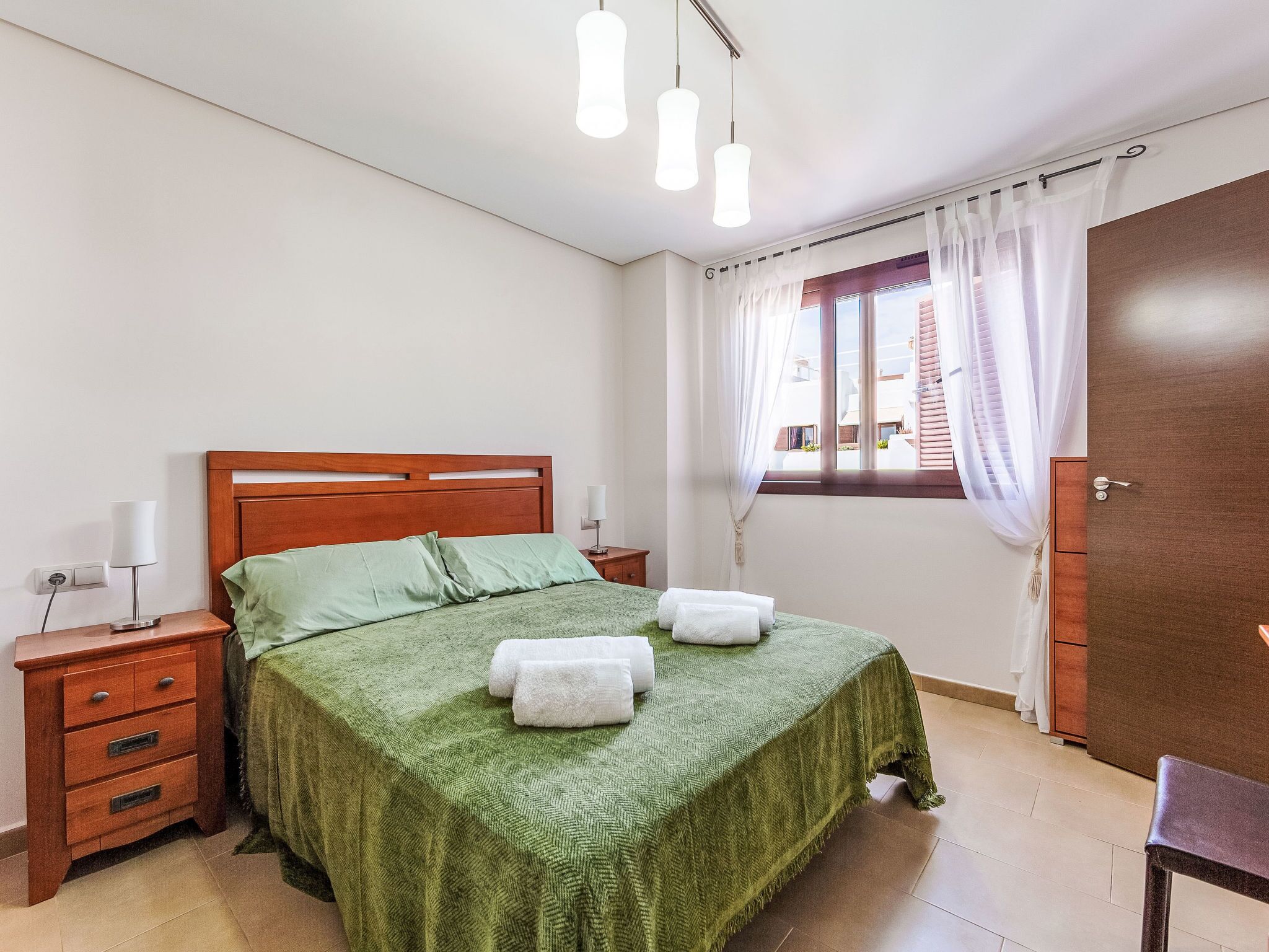 Een keurig opgemaakt slaapkamer met een bed, nachtkastjes, een lamp en een raam met gordijnen.