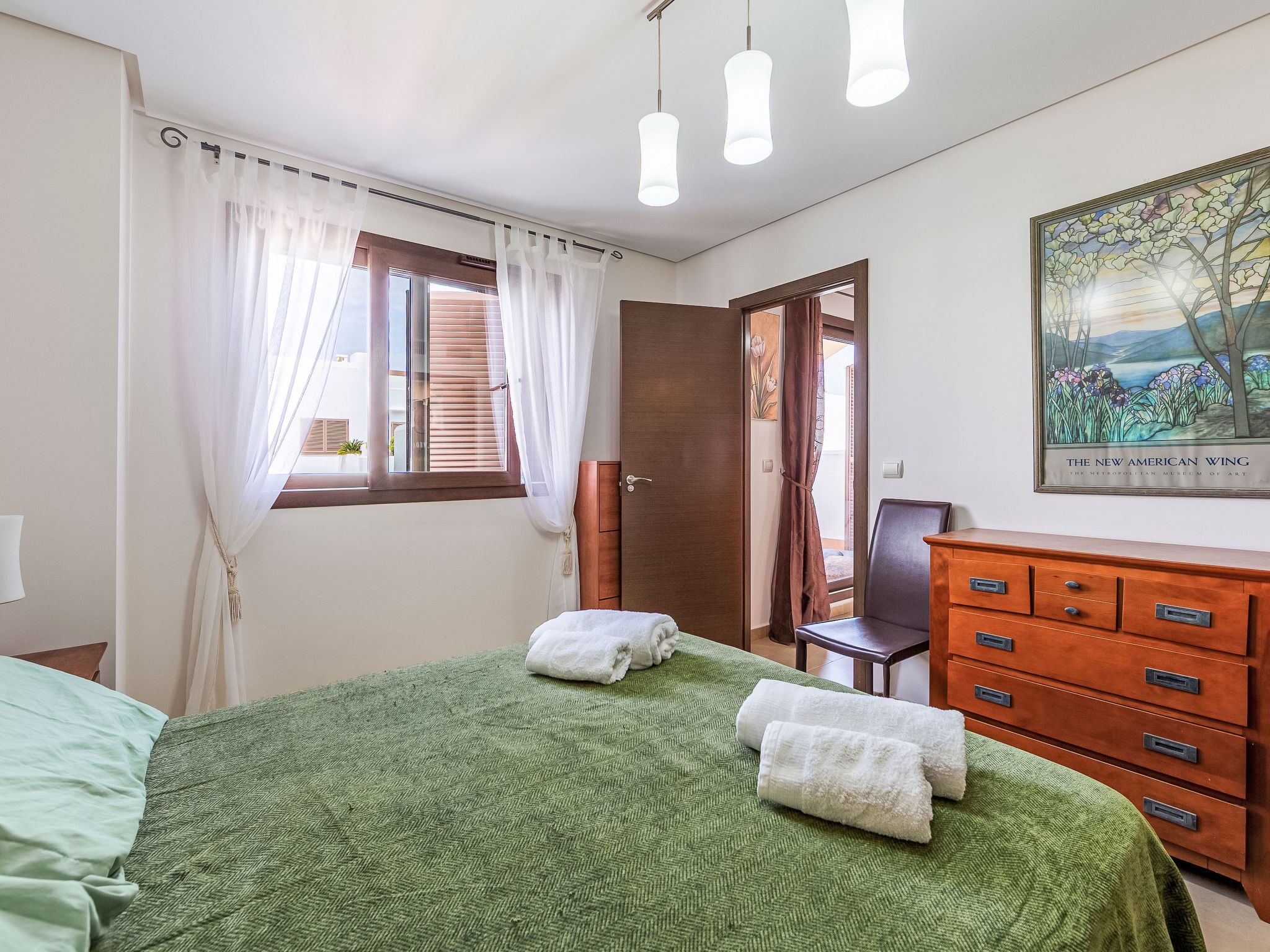A bedroom with a green bedspread, white towels, a wooden dresser, a chair, and a painting on the wall.