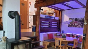 A cozy interior with a wood-burning stove, a dining area with wooden chairs and a table, and a wall-mounted shelf with decorative items. - Waldhaus57 Silbersee by Interhome (Frielendorf)