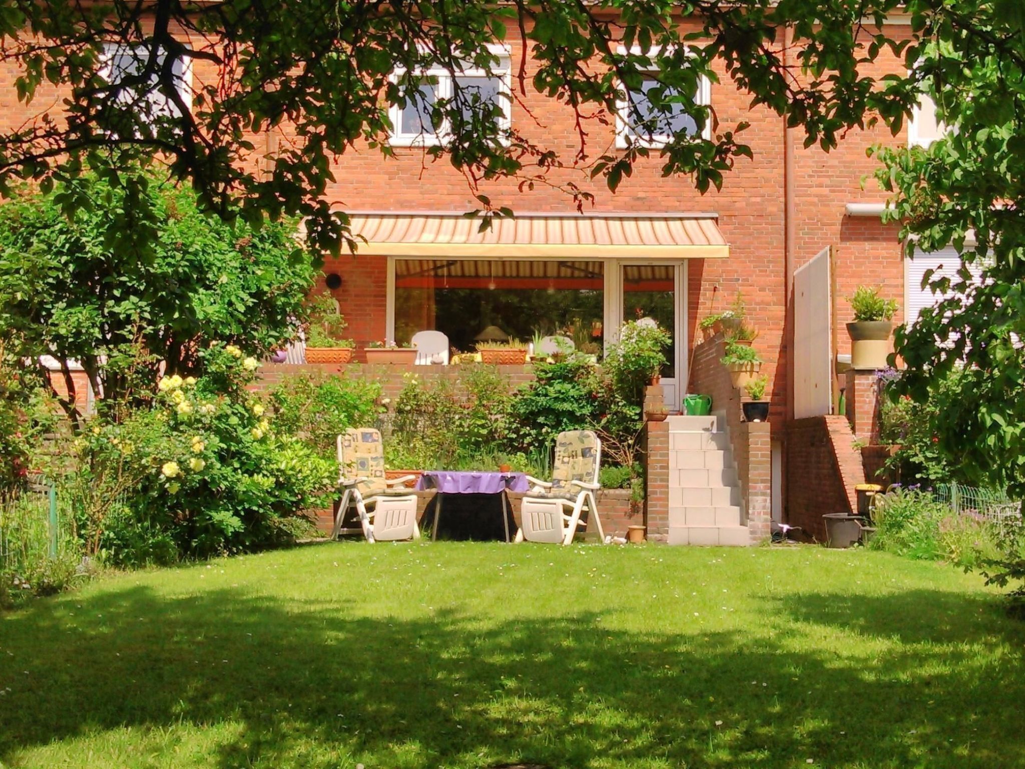 Eine Terrasse mit Tisch und Stühlen, umgeben von üppiger Bepflanzung und einem Ziegelgebäude mit überdachter Sitzgelegenheit.