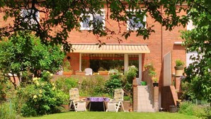 A patio area with a table and chairs, surrounded by lush greenery and a brick building with a covered seating area. - Wohnung in Altengroden mit Garten by Interhome (Wilhelmshaven)