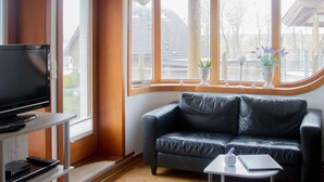 Un salon avec un canapé en cuir noir, un téléviseur posé sur un meuble en bois et une fenêtre donnant sur une maison et des arbres.