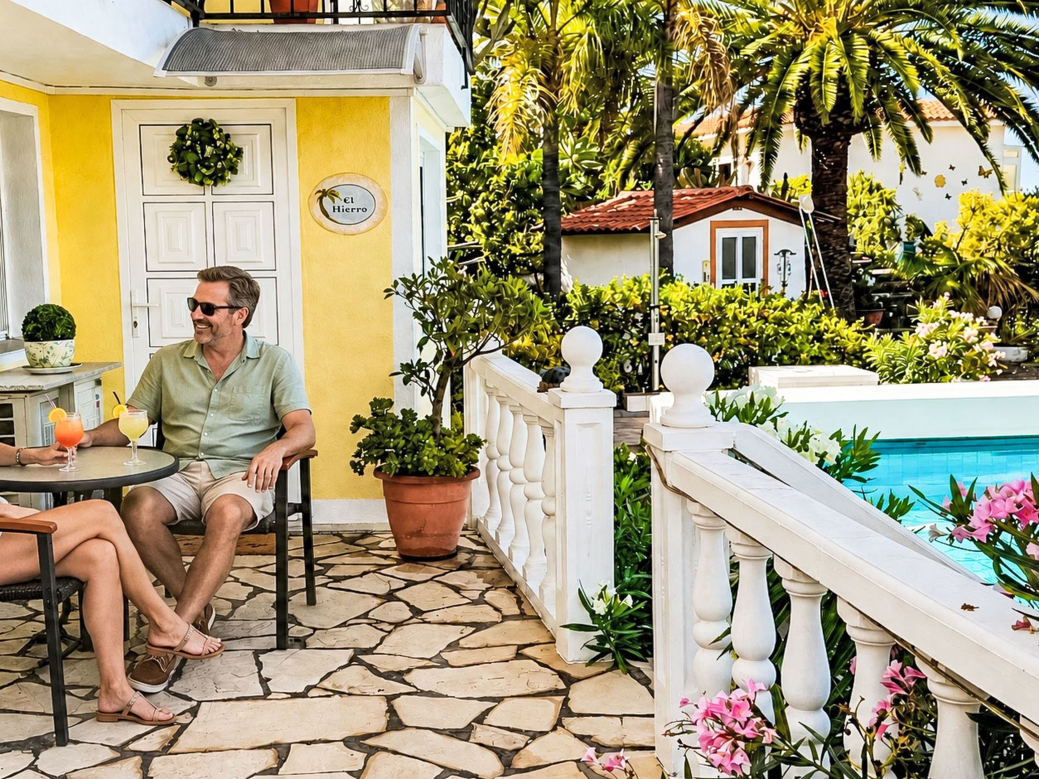 Ein Mann sitzt an einem Tisch im Freien mit Getränken, einem Topfpflanzen und Blick auf den Pool.