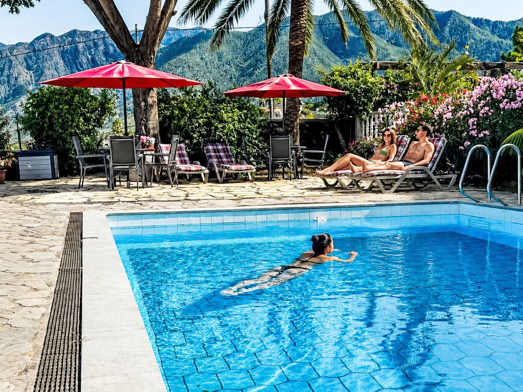 A swimming pool with lounge chairs and umbrellas, surrounded by greenery and mountains.