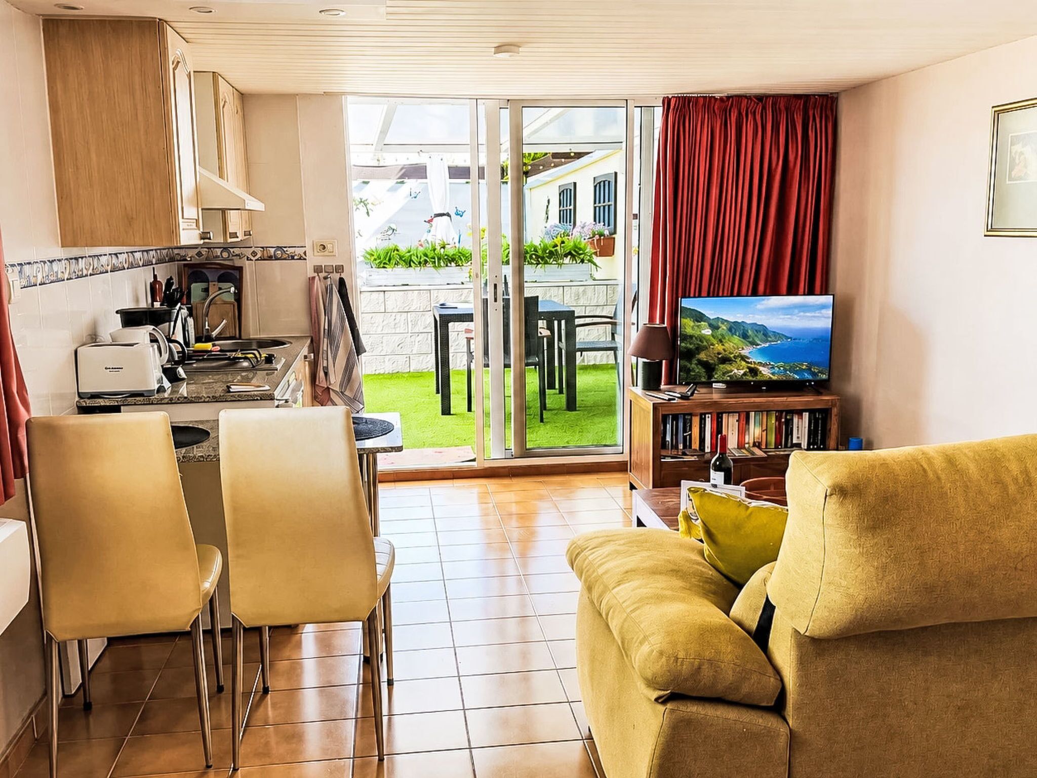 En moderne stue med sofa, spiseområde med stole, køkken med komfur og et fjernsyn, der viser et naturskønt udsyn.