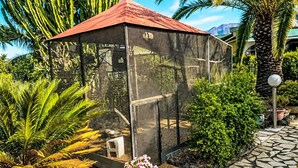 A covered outdoor area with a mesh roof, surrounded by tropical plants and a stone pathway. - La Gomera by Interhome (El Paso)