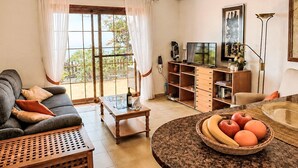 A living room with a sofa, coffee table, and a dining table with fruit. There is a TV, bookshelf, and a window with curtains. - La Gomera by Interhome (El Paso)