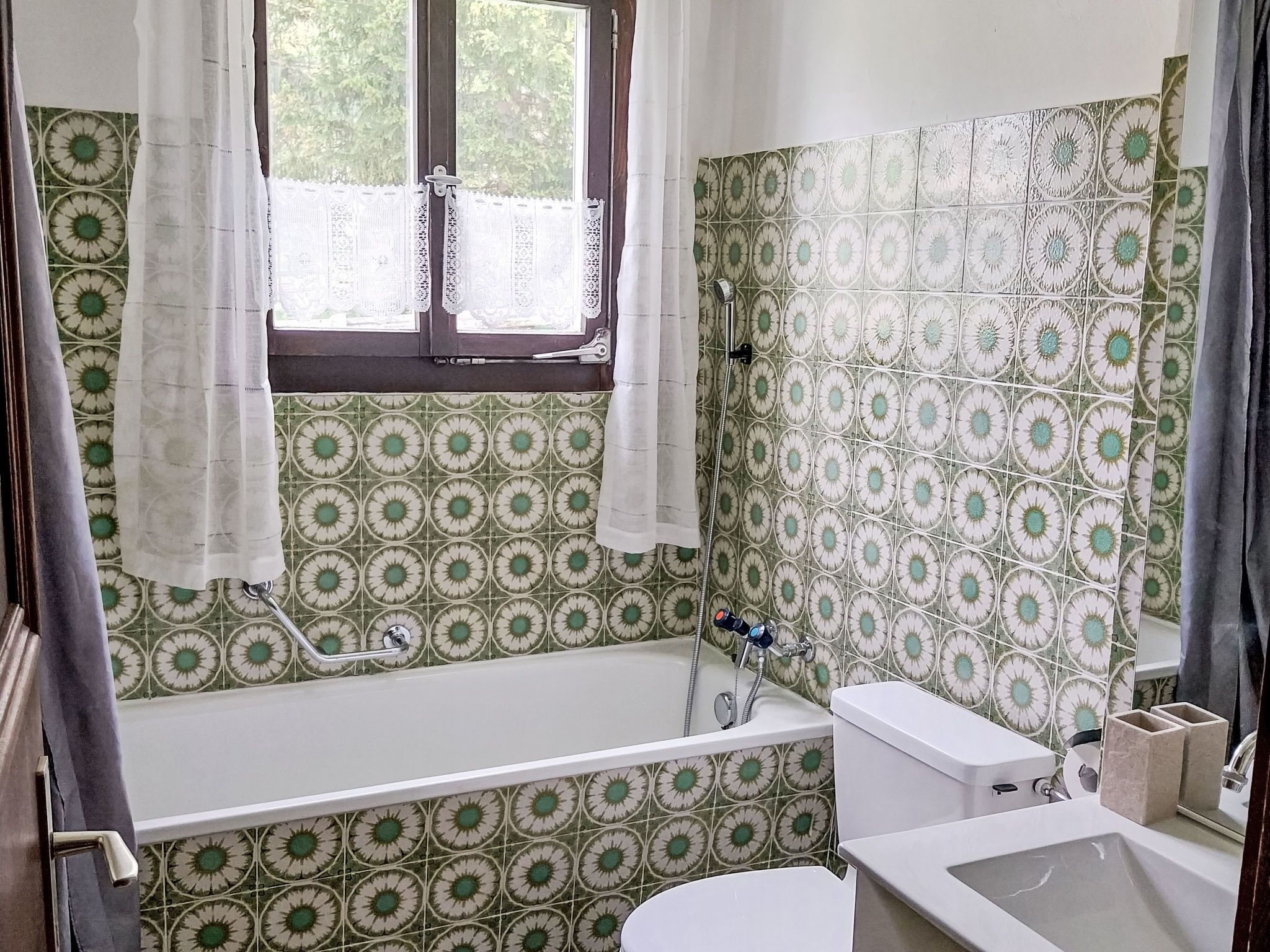 Un baño con bañera, inodoro y lavamanos, con azulejos estampados y un ventanal con cortinas.