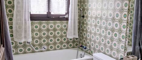 Un baño con bañera, inodoro y lavamanos, con azulejos estampados y un ventanal con cortinas.