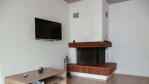 A living room with a flat-screen TV mounted on the wall, a wooden entertainment unit, and a fireplace with a tiled surround. - Chesa Blais by Interhome (Zuoz)